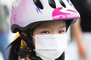 stänga upp ansikte friska flicka bär vit medicinsk ansikte mask. ljuv leende barn bär rosa hjälm för rida cykel. Lycklig unge med sport aktivitet i sommar eller vår tid. barn åldrig 5-6 år gammal foto