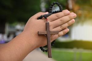 trä- korsa och trä- radband är hölls i händer av ung asiatisk katolik bön medan bön- i de tempel parkera område. foto