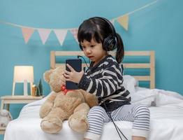 söt liten flicka sång med smartphone med teddy Björn i henne sovrum, Lycklig asiatisk barn liten flicka lyssnande de musik med hörlurar med teddy Björn på de säng, teknologi begrepp foto