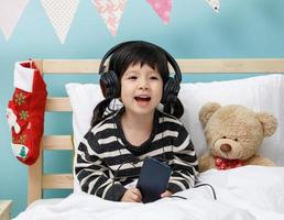 söt liten flicka sång med smartphone med teddy Björn i henne sovrum, Lycklig asiatisk barn liten flicka lyssnande de musik med hörlurar med teddy Björn på de säng, teknologi begrepp foto