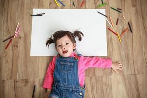 liten asiatisk flicka teckning i papper på golv inomhus, topp se av barn på golv foto