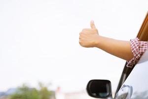 porträtt av Lycklig leende ung asiatisk man resande på de väg som visar tummen upp medan körning i hans bil. foto