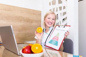 porträtt av ung leende kvinna dietist i de samråd rum. dietist skrivbord med friska frukt, juice och mätning tejp. dietist arbetssätt på diet planen. foto