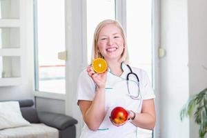 leende dietist i henne kontor, hon är innehav en frukt och som visar friska grönsaker och frukter, sjukvård och diet begrepp. kvinna dietist med frukt foto