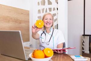 porträtt av ung leende kvinna dietist i de samråd rum. dietist skrivbord med friska frukt, juice och mätning tejp. dietist arbetssätt på diet planen. foto