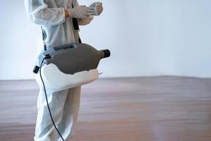professionell teknisk man i förebyggande kostym sprayer sterilisering lösning förbi elektrisk förbereder han själv innan spray maskin på de trä golv och vit bakgrund med studio ljus. foto