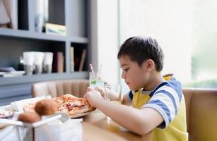 sida se porträtt unge äter Hem tillverkad pizza i de Kafé, Lycklig barn pojke bitande av stor skiva av färsk tillverkad pizza i de restaurang, familj Lycklig tid begrepp foto