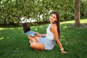 ung flicka Arbetar med en bärbar dator, Sammanträde på de gräsmatta. foto