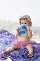 liten bebis flicka i rosa simning trunkar och panama Sammanträde på pläd på strand och dricka vatten från flaska. foto
