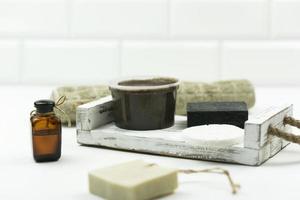 en choklad skrubba för kropp, en svart hand tillverkad tvål, en cooton handduk, en vit svamp och naturlig olja för ansikte stå på de vit trä- bricka i en modern badrum foto