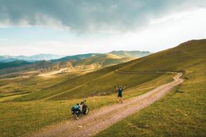 antenn inspirera se upphetsad glad caucasian manlig cyklist stående förbi röd touring cykel ser till naturskön bergen bakgrund och stå självsäker sorglös med händer upp foto