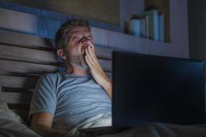 attraktiv trött och stressad workaholic man som arbetar sen natt utmattad på sängen upptagen med bärbar dator gäspande känsla sömnig och överansträngd i affärsprojekt tidsfristen stress koncept