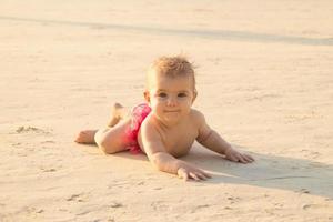 liten bebis flicka är liggande på en sandig strand nära till hav i solnedgång solljus. foto