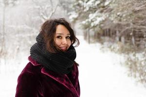skön leende kvinna med lång brun hår i en täcka från falsk päls och stor svart scarf på en bakgrund av vinter- skog. foto