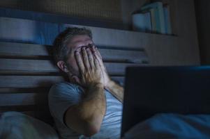 attraktiv trött och stressad workaholic man som arbetar sent på kvällen upptagen på sängen upptagen med bärbar dator känner sig sömnig och överarbetad i affärsprojekt deadline stresskoncept