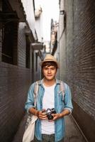 njuter resa. ung leende man med ryggsäck innehav kamera på asiatisk gata. foto
