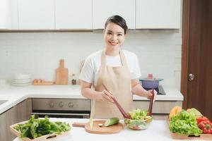 leende ung kvinna i förkläde stå i modern kök framställning sallad foto