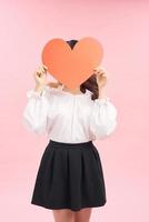 skön asiatisk flicka med en kort i de form av en hjärta i henne händer på en rosa bakgrund. foto