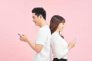 ung par är använder sig av smart telefoner och leende medan stående tillbaka till tillbaka på en rosa bakgrund foto