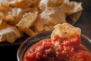 stänga upp hand håll chip dopp tomat salsa med tallrik med taco, nachos pommes frites och tomat dopp. kryddad nachos på rustik trä- bakgrund foto