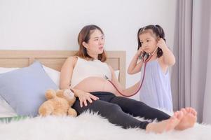ett asiatisk flicka användningar en stetoskop till känna henne mors mage till lyssna till henne bebis hjärtslag foto