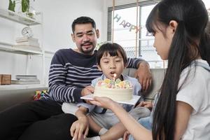 Lycklig asiatisk thai familj, ung son är överraskad med födelsedag kaka, slag ut ljus, och firar glädje fest med föräldrar och syster i levande rum tillsammans, välbefinnande inhemsk Hem särskild händelse. foto