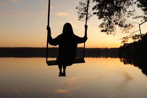 silhuett av en romantisk ung kvinna på en gunga över sjö på solnedgång. ung flicka resande Sammanträde på de gunga i skön natur, se på de sjö foto
