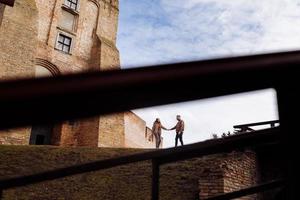 attraktiv ung par gående nära slott. fru och Make är gående innehav händer på bakgrund av gammal byggnad. två leende älskare. selektiv fokus foto