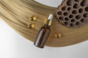 en hår vård olja eller serum i en brun dropper flaska och gyllene kapslar liggande på en blond hår strå, produkt marknadsföring attrapp foto