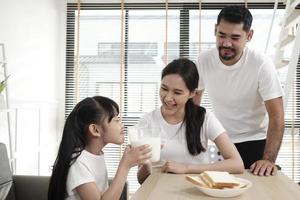 en friska asiatisk thai familj, en liten dotter, och ung föräldrar dryck färsk vit mjölk i glas och bröd glädje tillsammans på en dining tabell i morgon, wellness näring Hem frukost måltid livsstil. foto
