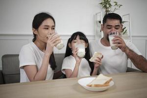 en friska asiatisk thai familj, en liten dotter, och ung föräldrar dryck färsk vit mjölk i glas och bröd glädje tillsammans på en dining tabell i morgon, wellness näring Hem frukost måltid livsstil. foto