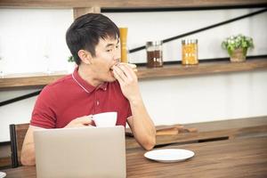 asiatisk leende ung man med avslappnad röd t-shirt njuter av att äta frukost, äta smörgås, ung man lagar mat och dryck i köksrummet i loftstil foto