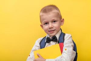 tillbaka till skola. en blond rolig pojke med en ryggsäck i en ljus skjorta på en gul bakgrund innehar böcker i hans händer och utseende på de kamera. kopia Plats. foto