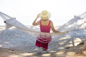 asiatisk kvinna kopplar av i hängmattan på den tropiska stranden, njut av sin frihet och friska luft, klädd i snygg hatt och kläder. glad leende turist i tropikerna i resesemester foto