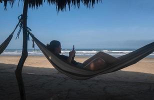 skön ung latinamerikan kvinna talande på henne cell telefon, vilar avslappnad i en hängmatta förbi de strand foto