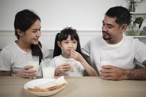 en friska asiatisk thai familj, en liten dotter, och ung föräldrar dryck färsk vit mjölk i glas och bröd glädje tillsammans på en dining tabell i morgon, wellness näring Hem frukost måltid livsstil. foto