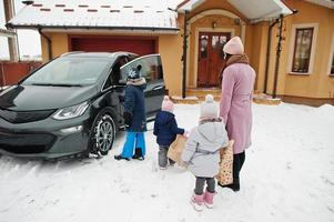 ung kvinna med barn håller ekoväskor och laddar elbil på gården till hennes hus. foto