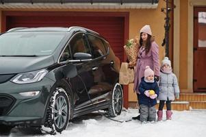 ung kvinna med barn håller ekoväskor och laddar elbil på gården till hennes hus. foto