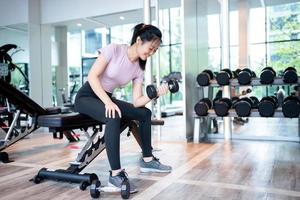 skön atletisk muskulös asiatisk kvinna pumps upp de muskler förbi ett ärm hissar hantel övning på bänk i kondition Gym. foto