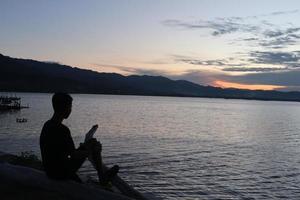 silhuett av en ung man stående förbi de sjö njuter de solnedgång. fredlig atmosfär i natur foto