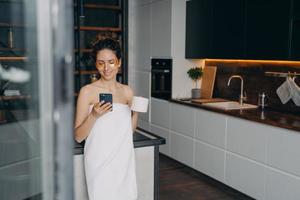 skön kvinna med under öga plåster använder sig av smartphone under hudvård rutin- efter morgon- dusch foto