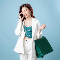halv kropp av självsäker skön asiatisk företag kvinna bär vit kostym talande på mobil telefon och innehav handla väska på isolerat blå bakgrund. tom skärm smartphone begrepp. foto
