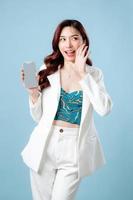 halv kropp av självsäker skön asiatisk företag kvinna bär vit kostym leende och som visar eller presenter mobil telefon Ansökan på hand på isolerat blå bakgrund. tom skärm smartphone begrepp foto