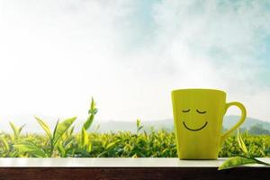 Lycklig och avslappning begrepp. en kopp av varm te med smiley ansikte på tabell i främre av grön te plantaion odla, berg med dimma som bakgrund foto