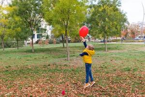 sorglös pojke spelar med röd ballong i höst parkera. foto