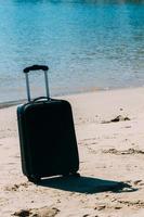 svart resa resväska på sandig strand med turkos hav bakgrund, sommar högtider begrepp foto