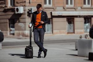 gående till flygplats terminal. självsäker affärsman resande gående på stad gator och dragande hans resväska dricka kaffe och tala på smartphone foto