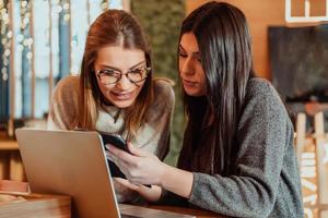 två ung företag kvinnor Sammanträde på tabell i Kafé. flicka visar kollega information på bärbar dator skärm. flicka använder sig av smartphone, bloggar. lagarbete, företag möte. foto