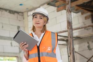 skön asiatisk kvinna arbetssätt på en konstruktion webbplats innehav en läsplatta medan bär en Hardhat och säkerhet väst med trötthet. foto