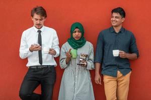 multietnisk grupp av tillfällig affärsmän använder sig av smartphone under en kaffe ha sönder från arbete i främre av de röd vägg utanför. foto